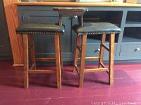 Two 25-inch tall stools with black cushioned seats and nailhead trim on legs of dark wood