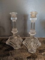 Pair of clear glass Tiffany & Co. candlesticks on a dark surface against a gray wall.