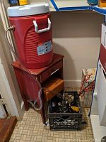 Red Coleman beverage cooler with white lid and attached clear plastic spigot, sitting on a small wooden stand with drawer and cupboard below, crate with keg taps and miscellaneous items on floor.