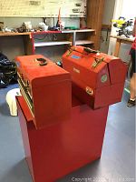 Photo showing two red metal tool boxes stacked on top of a large red tool cabinet, in a workspace with workbench and tools in background.