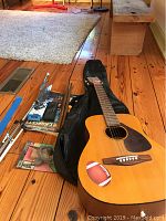 Yamaha FG Junior acoustic guitar lying on wooden floor with guitar bag underneath, assorted accessories including drum sticks, music books, and tuner visible beside it.