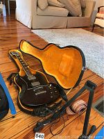 Black electric guitar inside an open hard case lined with orange plush fabric. The guitar has cables attached and is laying on a wooden floor indoors. The guitar has a double cutaway body style and chrome hardware.
