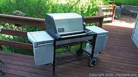 Full view of the Weber Genesis Gold gas grill sitting on a wooden deck, showing side shelves, metal frame, wheels, and grill body.