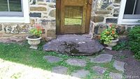 Wide angle photo showing both concrete planters placed on grass near a stone house entrance.