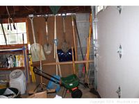 Wide view of the tools arranged in a garage corner, showing shovels, rakes, spreader, broom, and digging tools.