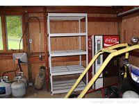 Full view of white plastic shelving unit in garage setting with four shelves, near Coca-Cola vending machines and other garage items.