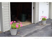Two of the gray plastic planters with pink geraniums positioned outside a garage door with part of the driveway visible.
