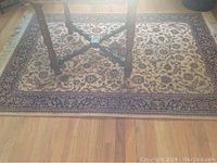Photo showing one of the Kurdish wool rugs under a table, beige with intricate floral design and navy border