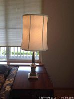 Side view of one brass pedestal base table lamp with lit off-white linen shade on wooden side table next to floral patterned couch.