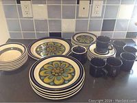 Full set show on countertop, including plates, bowls, mugs, sugar bowl with blue and olive floral pattern and solid blue mugs.