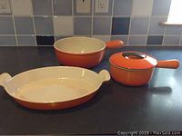 Photo showing three orange Le Creuset cast iron cookware pieces on a kitchen counter with tile backsplash. Includes a shallow rectangular baking dish, a small saucepan with lid, and a medium saucepan without lid.