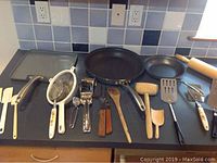 A variety of kitchen utensils including spatulas, wooden spoons, a rolling pin, and two frying pans on a kitchen counter with a tile backsplash.