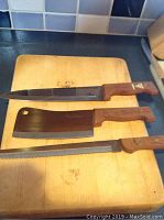 Three knives on cutting board: long carving knife, large cleaver, serrated bread knife, all wood handled