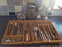Photo showing a wooden tray with various stainless steel flatware including forks, knives, spoons, serving utensils, a box cheese grater, salt and pepper shakers, and a small container with lid.