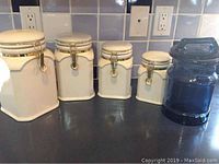 Four white ceramic canisters with gold-tone clasps arranged side by side on counter.