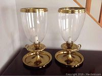 Photo showing two tall brass and glass candle holders placed side by side on a flat surface, highlighting the design and condition.