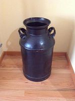 Front view of the black vintage metal milk can standing on floor, showing its round base, smooth cylindrical body, two handles near the top, and open mouth rim.