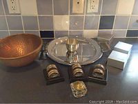 Overall shot showing copper bowl on left, pewter serving tray with wooden handles in center, small pewter bowl on tray, 3 pairs of silver-plated napkin rings displayed in front, and glass ink well with brass lid in front center.