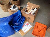 Overview of boxes containing tarps and painting supplies including a blue tarp, orange paint tray, and cardboard boxes.