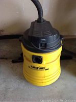 Front angle showing yellow Genie wet/dry vacuum with hose attached, control switch on the front, and round yellow base.