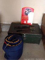 Green military trunk with items resting on top and blue rolled sleeping bag beside it