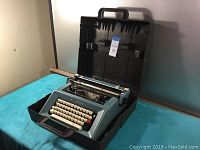 Blue Olivetti typewriter in original hard black carrying case with handle, showing keyboard, platen and ribbon mechanism.