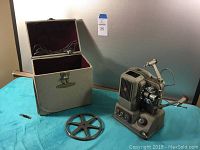 Full view of Elmo E-80 projector, metal carrying case with open lid showing fabric interior and cords, and a metal film reel laid flat on blue cloth surface.