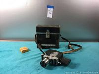 Photo of the Keystone K-808 Zoom projector, black carrying case with strap and latch, and a small film box on a blue cloth surface.