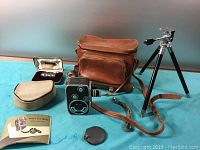 Photo showing Bolex C8 camera, leather carrying case, tripod, lens cases, manual, and accessories on a blue surface.