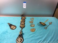 Full view of the horse brass set on leather straps laid flat with a cluster of 5 additional loose brass plaques and two brass serving utensils on the right.