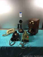Full view of the five items on blue display table including 2 rotary phones, push-button phone, wooden phone box, and replica candlestick phone radio.