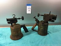 Photo showing two antique soldering blow torches side by side on a blue cloth background, one with 'Shapleighs' label, the other with wooden handle