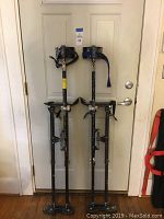 Front view of two adjustable drywall stilts against a door showing overall height, metal frames with paint marks, foot platforms, and ankle straps (one black and one blue).