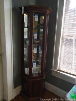 Front view of the tall wooden curio cabinet with glass panels, lighting inside, and visible multiple shelves, photographed against a corner wall near a window.