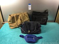 Four bags including two ladies handbags, a laptop bag, and a blue fanny pack, arranged on a turquoise surface with a scoreboard background.