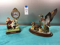 Both figurines together on blue cloth background: ceramic clock with children figurines and porcelain duck family figurine
