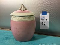 Pink ceramic jar with matching lid, showing embossed pale green band and sculpted twig handle on lid.