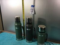 Three Stanley thermoses of different sizes and colors on a blue cloth with a tape measure behind them showing size.