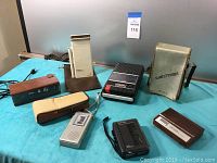 Full view of seven vintage electronic devices on blue table including Sony Watchman TV in case, Radio Shack cassette recorder, Norelco fabric steamer, Panasonic tape recorder, and four other audio devices.
