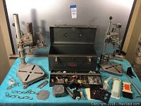 Photo showing the vintage Craftsman metal toolbox open with miscellaneous mechanical parts inside and two metal drill press stands placed on either side on a light blue cloth.