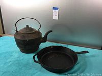 Photo showing cast iron skillet and iron kettle together against gray background on blue cloth.
