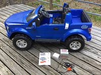 Side view of the blue Fisher Price Power Wheels F150 electric ride-on truck showing two seats, remote, battery charger, and manuals laid out beside it.