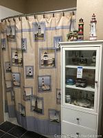 Shower curtain with lighthouse and seashell themed patterns hanging on bronze-colored rings on a curved shower rod. Three decorative lighthouse figurines in different styles and colors on top of a white cabinet beside the shower. Cabinet not included in lot.