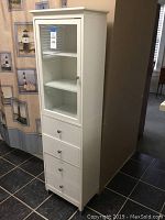 Front view of tall white bathroom organizer stand showing glass door with shelves and drawers below.