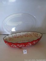 Front view of the red and cream oval stoneware baker with intricate floral lace pattern and clear lid placed behind it.