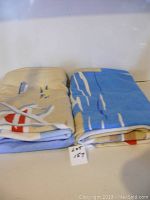 Two folded beach towels on a shelf, one beige with sailboat design, the other blue with wave pattern, labeled lot 157.