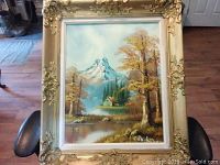 Full view of framed oil painting depicting a mountain landscape with a cabin and trees with autumn foliage.