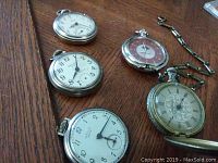 Top view of five pocket watches arranged on wood surface, showing white-faced Westclox watches and Harley Davidson with red face and metal chain.