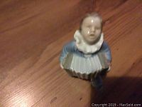 Front view of a blue-clad clown figurine sitting and playing an accordion, showing details of the face and ruffled collar.