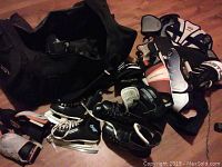 Large black hockey bag open with multiple hockey gear items including two pairs of size 8 skates, gloves, shin guards, and elbow pads visible around the bag